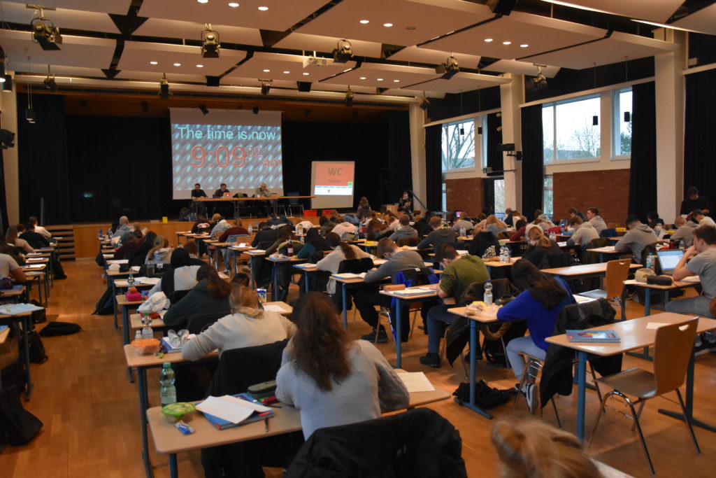 Prüflinge sitzen an Einzeltischen in einem großen Saal.
