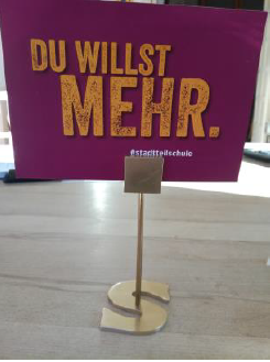 Selbst gebauter Kartenhalter aus Metall vor einem Schild „Du willst mehr.“