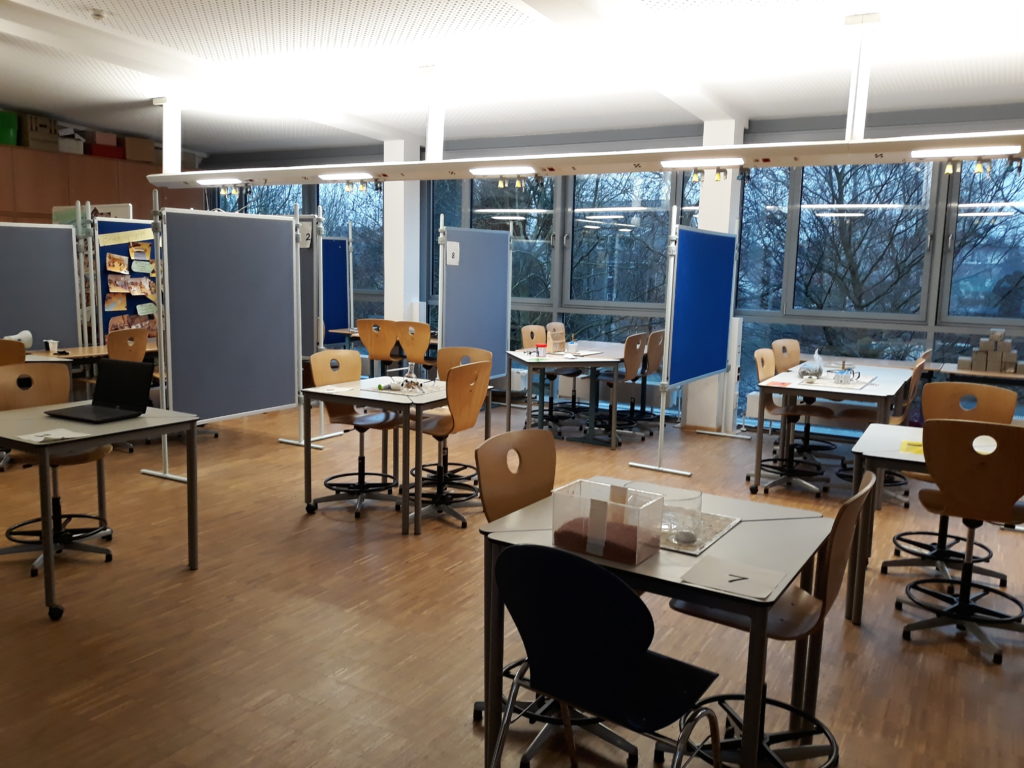 Klassenraum mit Tischgruppen, Stühlen und großen Fenstern.