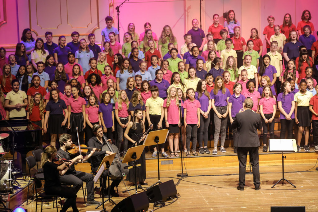 Großer Chor in bunten T-Shirts mit Orchester und Dirigent auf der Bühne.