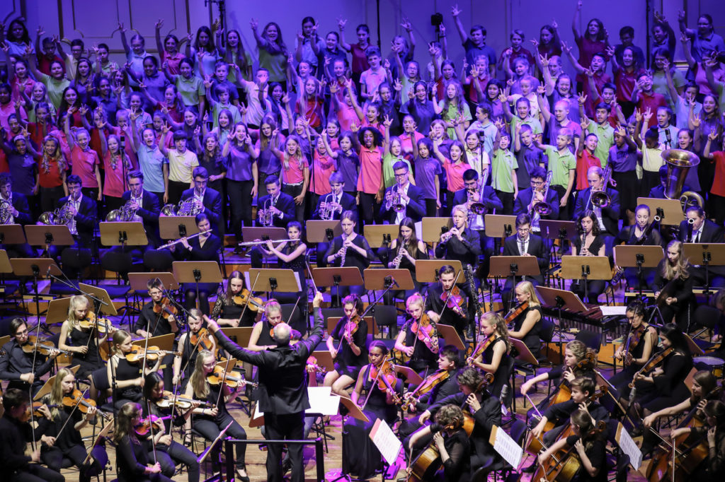 Hunderte Sängerinnen und Sänger und ein Orchester in einem großen Konzertsaal.