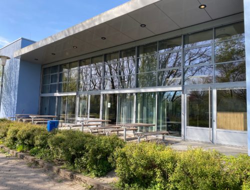 Glasfront der Mensa mit Terrasse und Sitzplätzen draußen.
