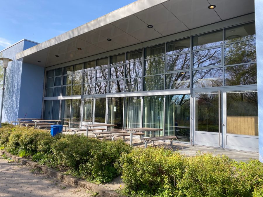 Glasfront der Mensa mit Terrasse und Sitzplätzen draußen.