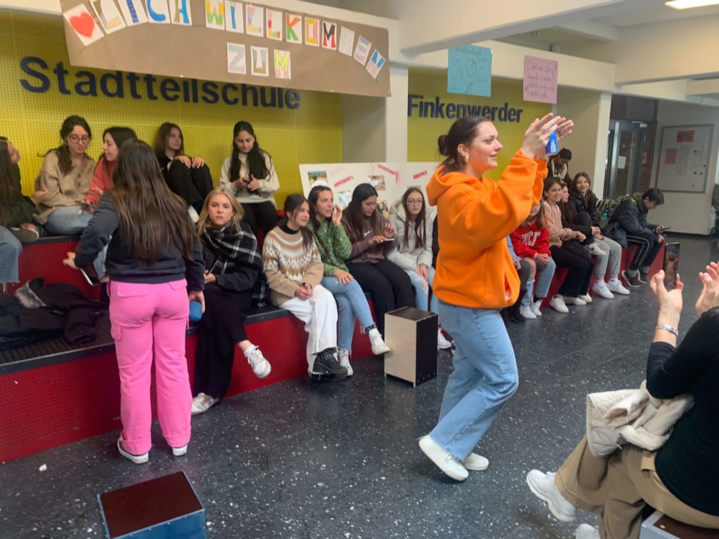 Schülerinnen und Schüler sitzen in der Pausenhalle, eine Schülerin tanzt.