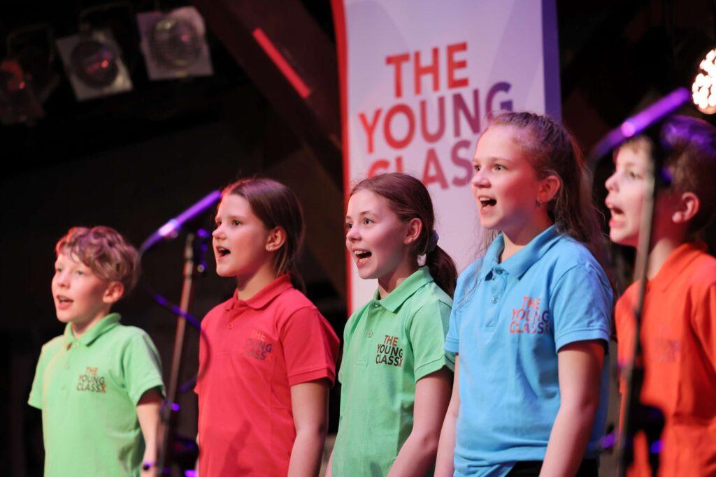 Kinder in bunten T-Shirts singen auf der Bühne der Fabrik.