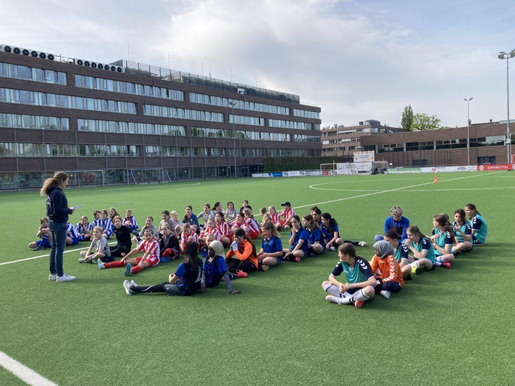 Schülerinnen und Schüler sitzen im Kreis auf einem Sportplatz.