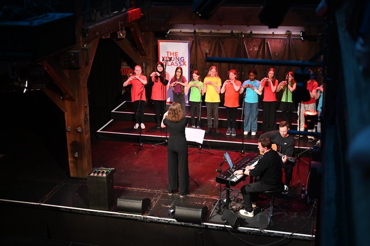 Chor in bunten T-Shirts auf der Bühne der Fabrik, vorne eine Band.