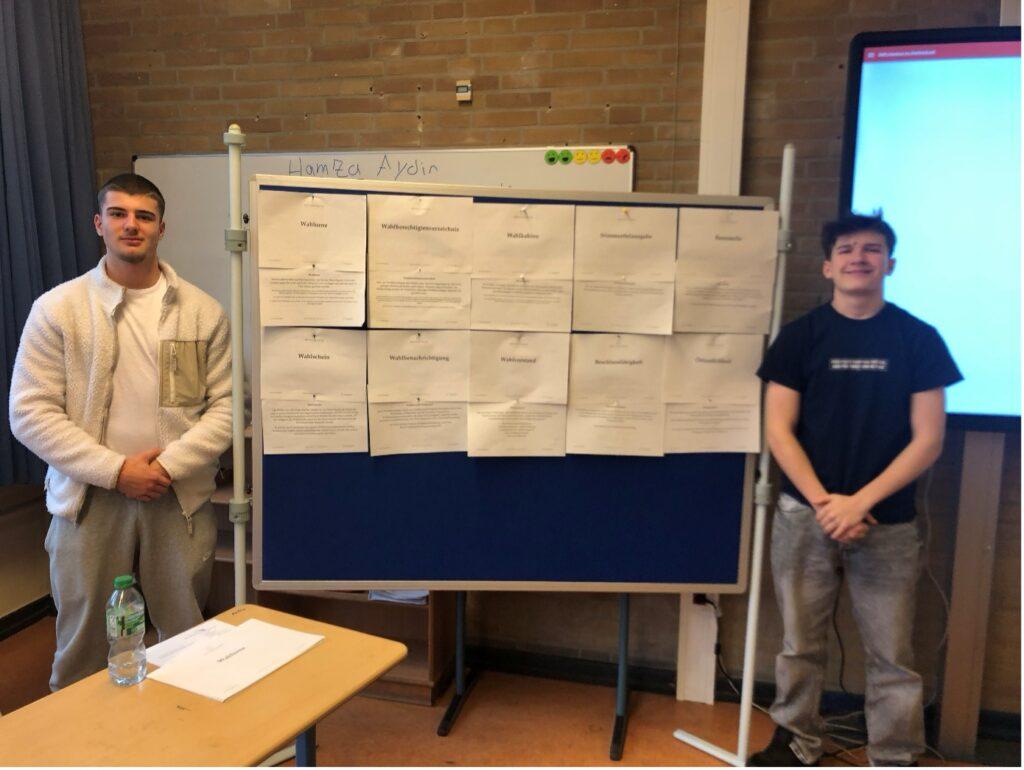 Zwei Workshopleiter vor einer Pinnwand mit Plakaten.